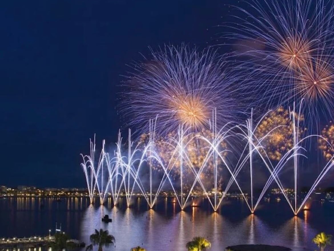 Un feu d’artifice du Festival d’Art Pyrotechnique éclatant au-dessus de la baie de Cannes, reflété dans l’eau, avec les palmiers du front de mer au premier plan. — Antibes · Juan-les-Pins