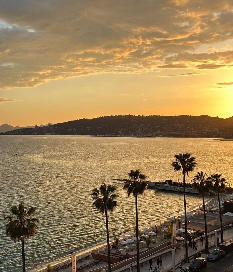 Le coucher de soleil sur la baie de Juan-les-Pins, vu depuis la terrasse : les palmiers, la plage et le front de mer. — Palais Juan, Juan-les-Pins