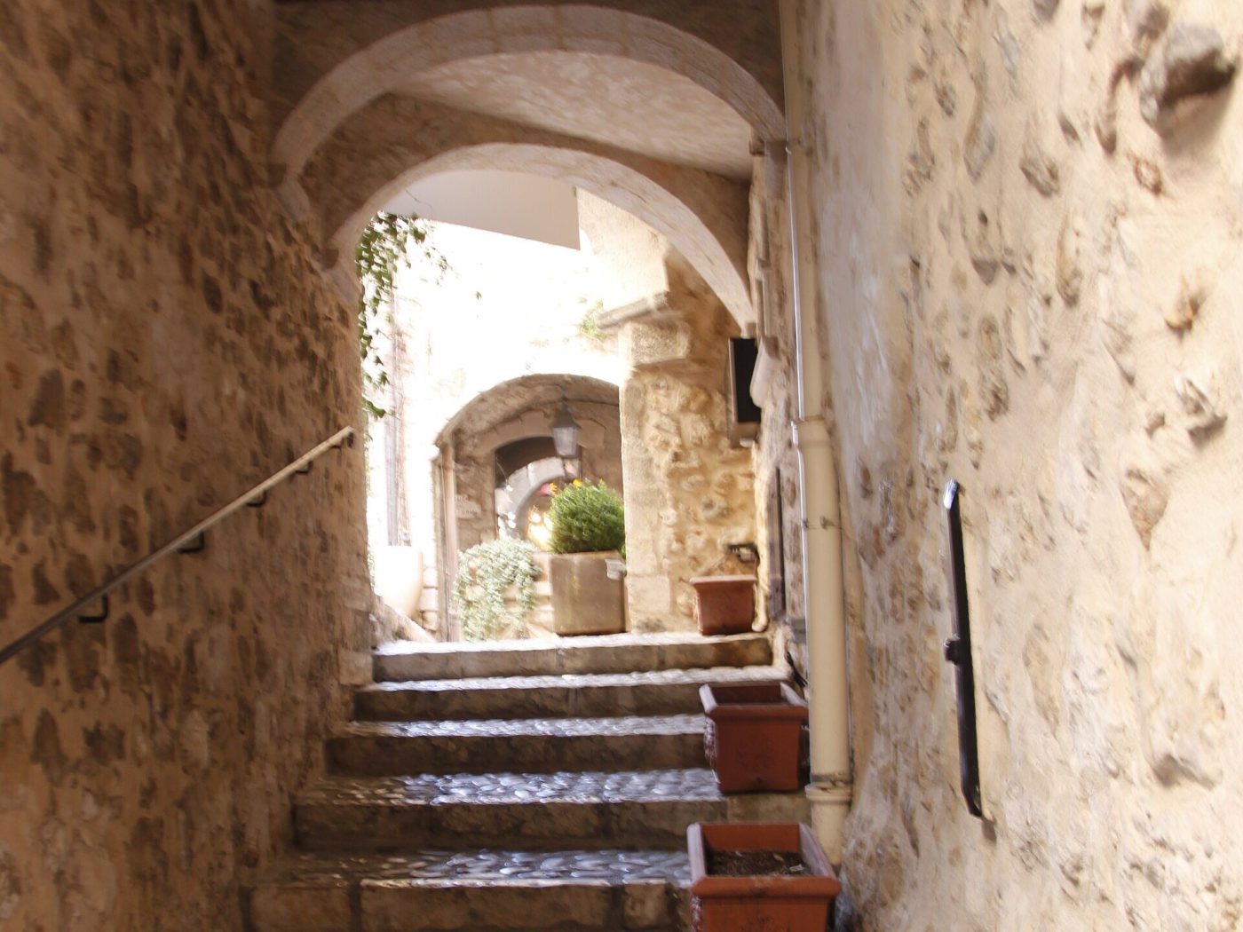 Un escalier de pierre sous les voûtes d’une ruelle de Saint-Paul-de-Vence, bordé de pots en terre cuite. — Antibes · Juan-les-Pins